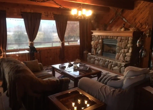Cozy living room with stone fireplace, wooden walls, and large windows overlooking a scenic view. Warm lighting enhances the ambiance.
