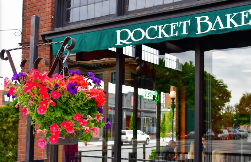 A storefront with a green awning reading "Rocket Bakery" and vibrant flower baskets hanging outside.