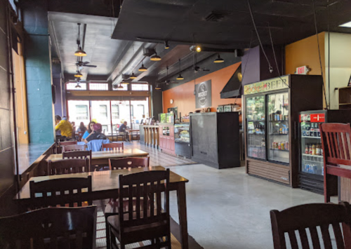 Interior of a casual restaurant with wooden tables, chairs, and a beverage fridge, featuring a bright, welcoming atmosphere.