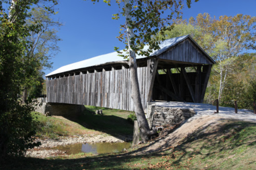 A wooden covered bridge spans a dry creek, surrounded by trees and a clear blue sky.