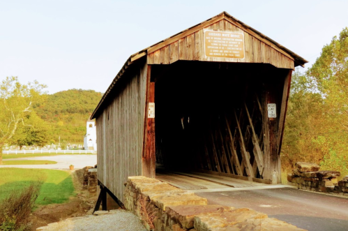 A rustic wooden covered bridge with a stone foundation, surrounded by greenery and a clear sky.