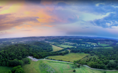 Aerial view of a lush green landscape under a colorful sunset sky, with rolling hills and scattered trees.
