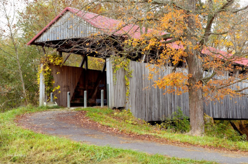 A rustic covered bridge with a red roof, surrounded by autumn foliage and a winding path.