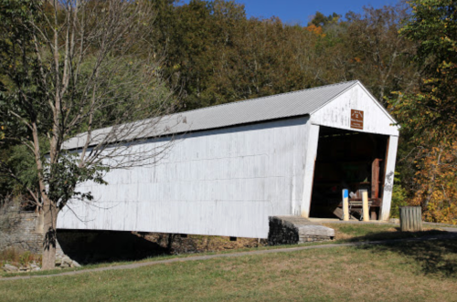 A white covered bridge spans a small stream, surrounded by trees and greenery on a sunny day.