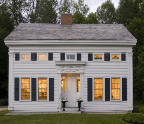 A white historic house with blue shutters, large windows, and a central door, surrounded by greenery.