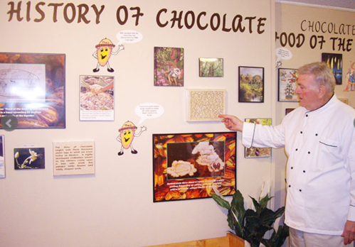 A chef points to a display about the history of chocolate, featuring images and text on a museum wall.