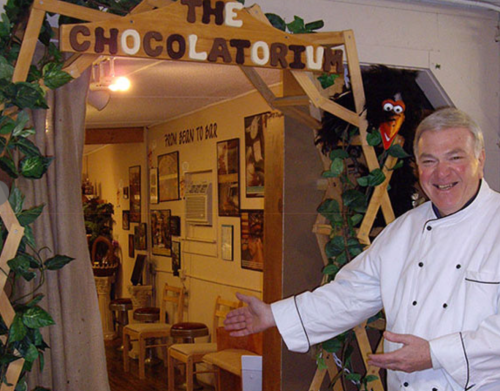 A smiling chef gestures towards an entrance labeled "The Chocolatorium," surrounded by greenery and decor.