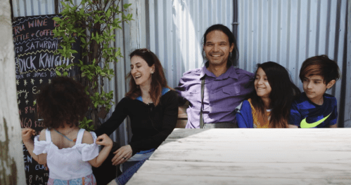 A family of five smiles together at a café, with a chalkboard menu in the background and greenery nearby.