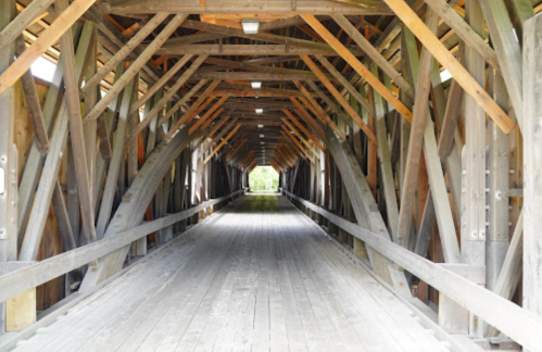 A wooden covered bridge interior with exposed beams and a wooden floor, leading to a bright opening at the end.