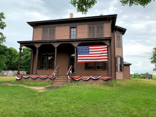 A historic brown house with a large American flag and red, white, and blue bunting on the porch, surrounded by green grass.