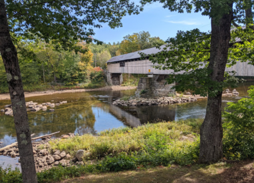A covered bridge spans a calm river, surrounded by trees and autumn foliage under a clear blue sky.