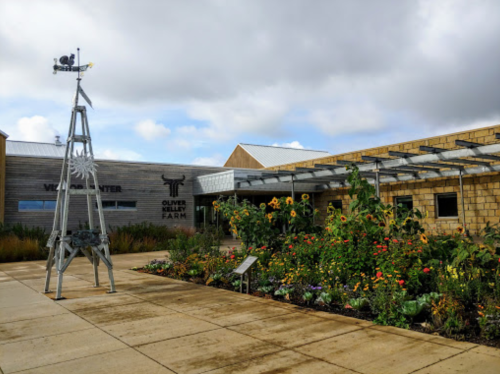A modern farm building with a windmill and vibrant flower beds under a cloudy sky.
