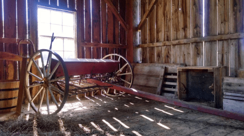 An old wooden barn interior featuring a vintage cart and wooden crates, illuminated by sunlight through a window.