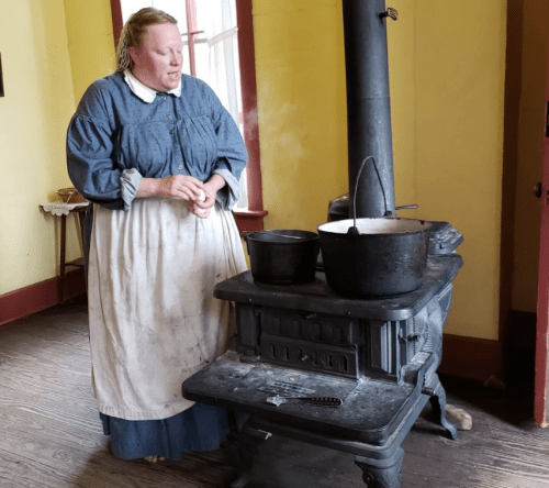A woman in historical attire stands by a wood stove, cooking in two pots, with steam rising in a rustic kitchen.