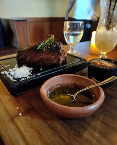 A sizzling steak on a hot stone, accompanied by a small bowl of sauce and a side dish, with a glass of water in the background.