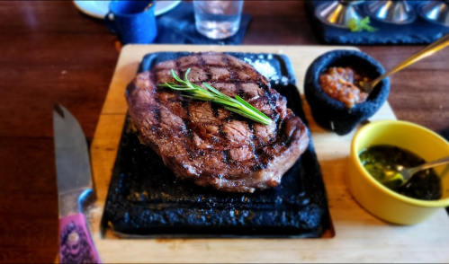 A grilled steak on a wooden board, garnished with rosemary, accompanied by sauces in small bowls.