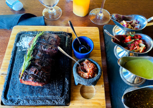 A grilled steak on a hot stone, accompanied by salsas, sauces, and fresh toppings on a wooden table.