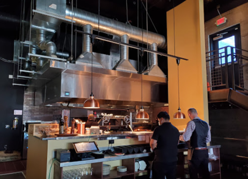 A modern restaurant kitchen with metal ventilation, two chefs working, and a stylish interior design.