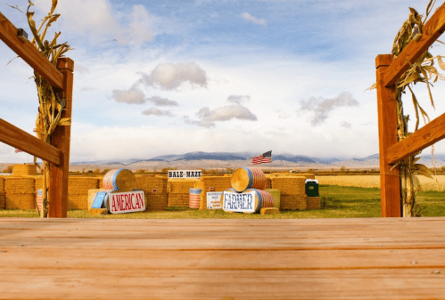 A wooden deck overlooks a farm scene with hay bales, signs, and a distant mountain under a cloudy sky.