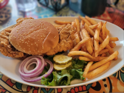 A plate with a fried chicken sandwich, fries, lettuce, pickles, and red onion on a colorful tablecloth.