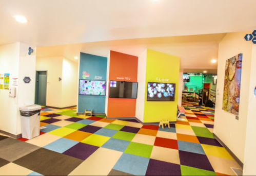 Colorful hallway with interactive screens and a vibrant carpet, leading to different rooms in a playful environment.