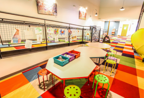 Colorful classroom with octagonal tables, chairs, and vibrant carpet, featuring art displays on the walls.