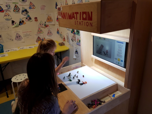 Two children create stop-motion animations at a wooden animation station with a screen and figurines.