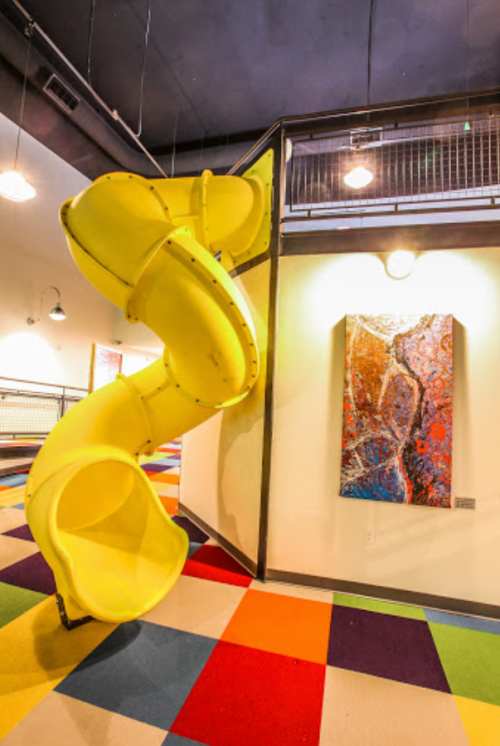 A bright yellow slide spirals down from a wall in a colorful play area with abstract art nearby.