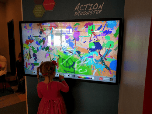 A child in a pink dress interacts with a colorful digital art display, creating vibrant splashes of paint.