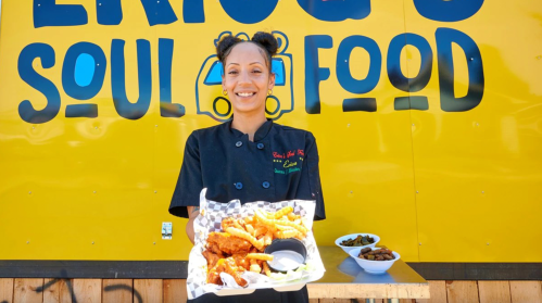 A smiling chef holds a plate of fried chicken and fries in front of a bright yellow food truck sign.