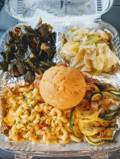 A takeout container with macaroni and cheese, cornbread, sautéed zucchini, collard greens, and cabbage.