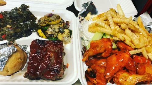 A takeout container with collard greens, grilled vegetables, meatloaf with sauce, and crispy fries with chicken wings.