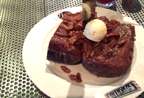 A plate of rich chocolate cake topped with a scoop of ice cream and pecans, served on a metal table.