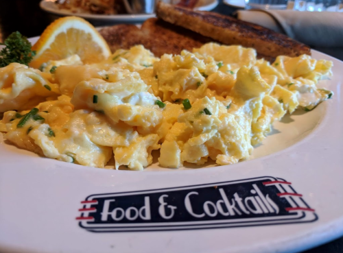 A plate of scrambled eggs with herbs, a lemon wedge, and toast, with a restaurant logo at the bottom.