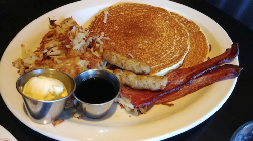 A plate of pancakes, crispy bacon, sausage links, and hash browns, served with butter and syrup.