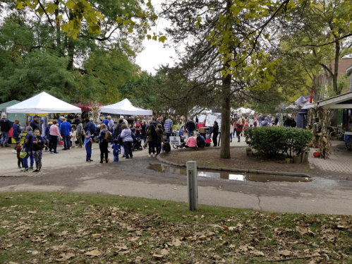 A bustling outdoor market with tents, people browsing, and autumn leaves scattered on the ground.