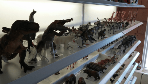 A display of various animal figurines, including dinosaurs, horses, zebras, and giraffes, on white shelves.