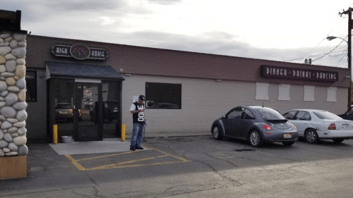 A man stands outside a building with a sign for "Dinner, Drinks, Dancing" next to parked cars.