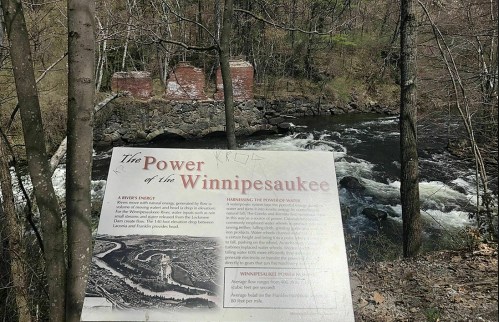 A sign about the power of the Winnipesaukee River, with a river and old stone structures in the background, surrounded by trees.