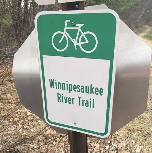 Sign for the Winnipesaukee River Trail featuring a bicycle symbol, set against a natural background.