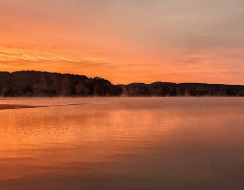 A serene lake at sunset, with mist rising and vibrant orange and pink hues reflecting on the water.