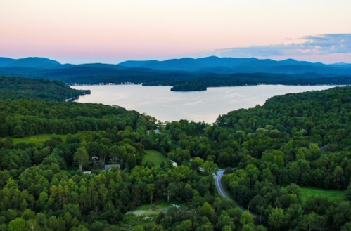 Aerial view of a serene lake surrounded by lush green forests and distant mountains at sunset.