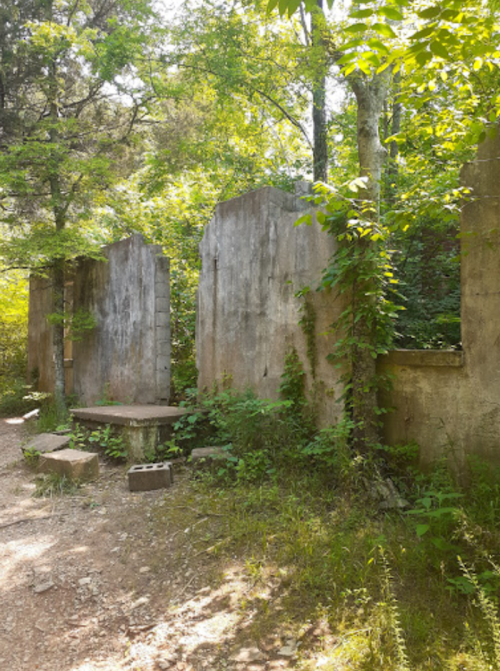 Ruins of concrete walls covered in greenery, surrounded by trees and a gravel path.