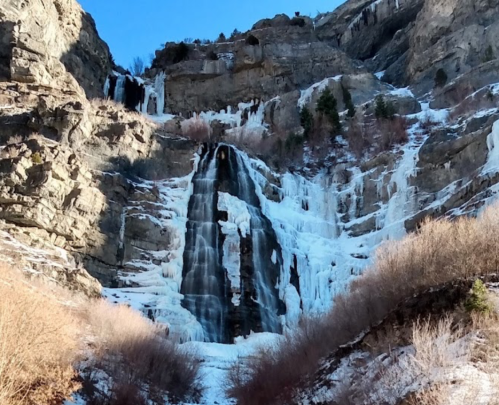 A frozen waterfall cascading down a rocky cliff, surrounded by snow and ice under a clear blue sky.