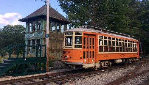 An orange vintage streetcar parked near a historic signal tower surrounded by trees and a gravel path.