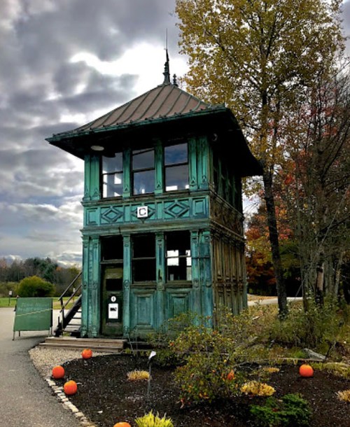 A vintage green tower with a peaked roof, surrounded by trees and pumpkins, under a cloudy sky.
