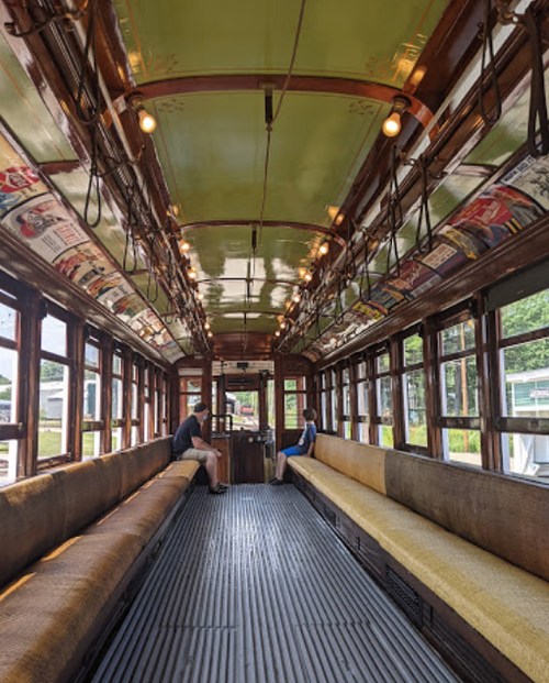 Interior of a vintage streetcar with wooden benches, green ceiling, and two passengers sitting at opposite ends.
