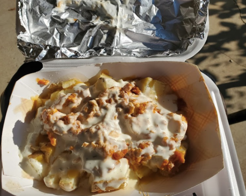A takeout container filled with fries topped with creamy sauce and crumbled toppings, sitting on a surface.