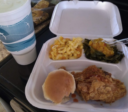 A takeout container with fried chicken, macaroni and cheese, greens, a biscuit, and a large drink.