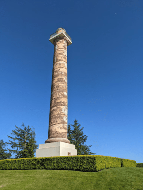 A tall, decorative stone column stands against a clear blue sky, surrounded by neatly trimmed green hedges.